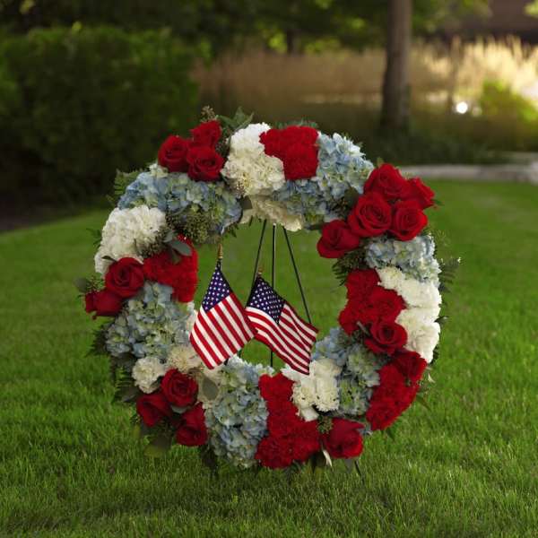 Floral wreath with red roses, white carnations, blue hydrangeas, and two small American flags