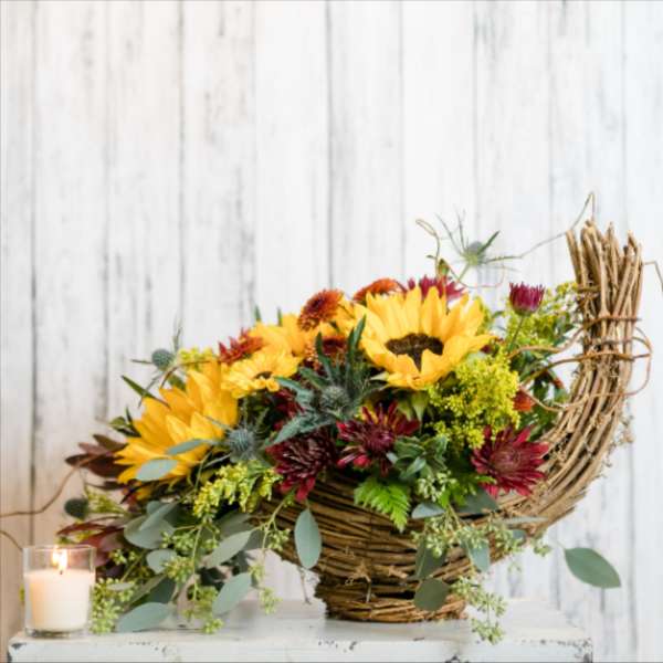 Basket arrangement with sunflowers and burgundy flowers beside a lit candle