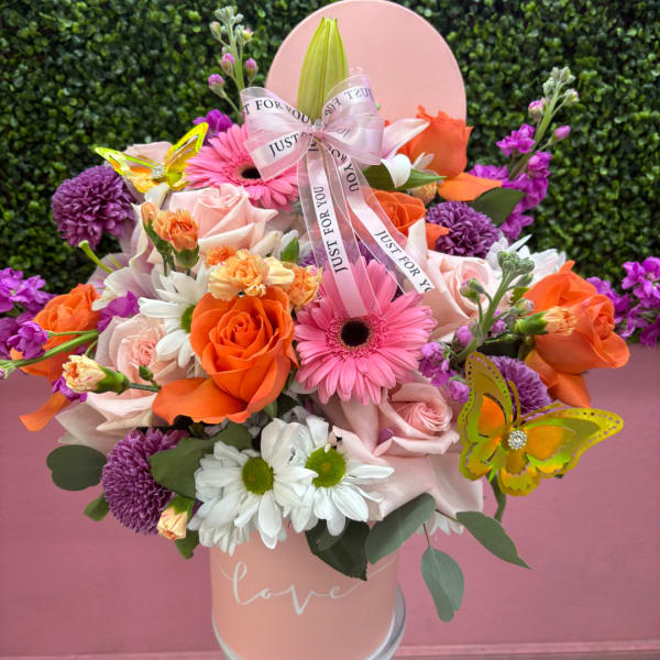 Pink and orange mixed flower arrangement in a round box with ribbon and butterfly accents