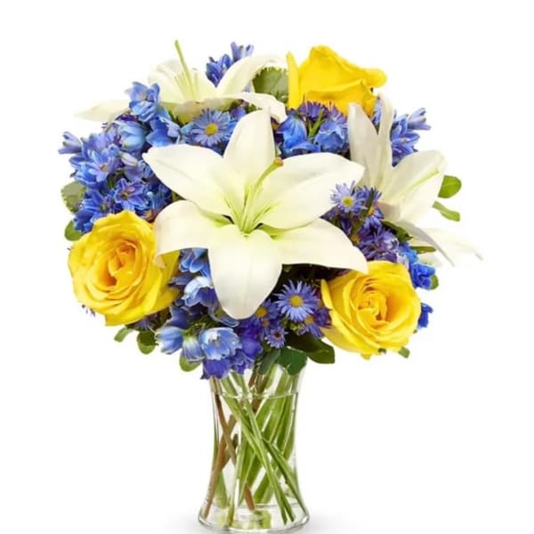 Bouquet of white lilies, yellow roses, and blue flowers in a clear glass vase