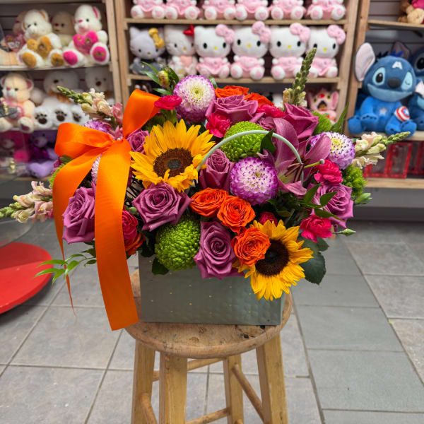 Bright mixed bouquet with sunflowers, purple roses, and an orange ribbon in a square box