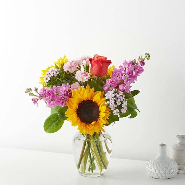 Mixed bouquet with a sunflower, rose, and pink blooms in a clear glass vase