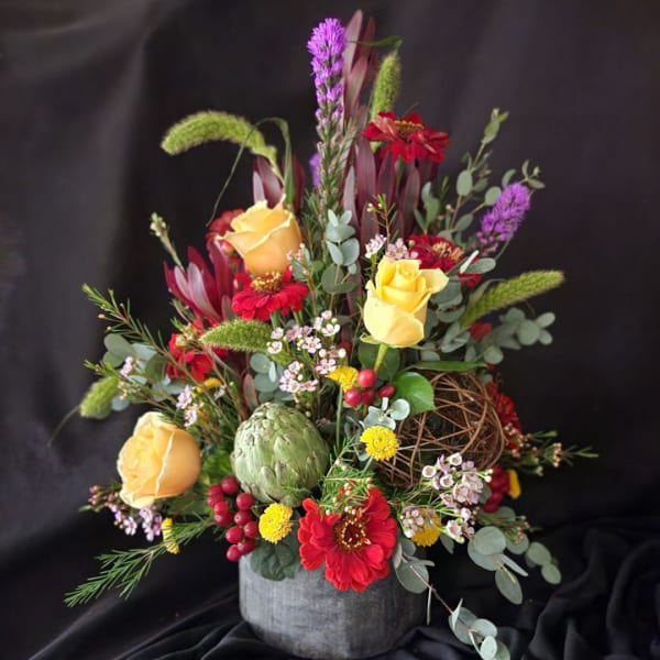 Mixed floral arrangement with yellow roses, red blooms, and a green artichoke in a gray vase