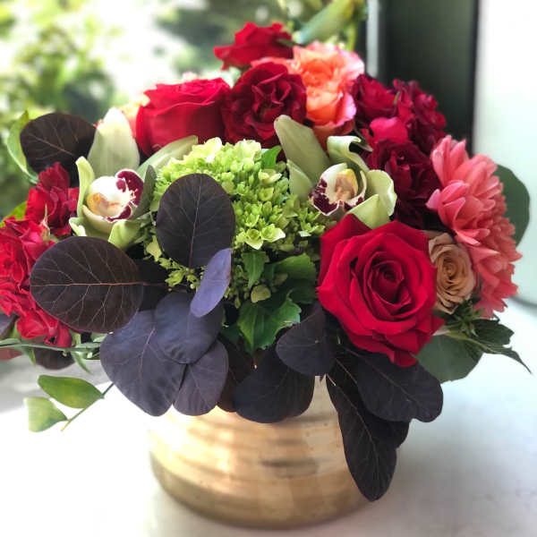 Low arrangement of red roses, green hydrangea, orchids, and mixed blooms in a gold vase