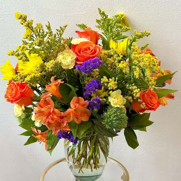 Mixed bouquet of orange roses, yellow blooms, and purple flowers in a glass vase