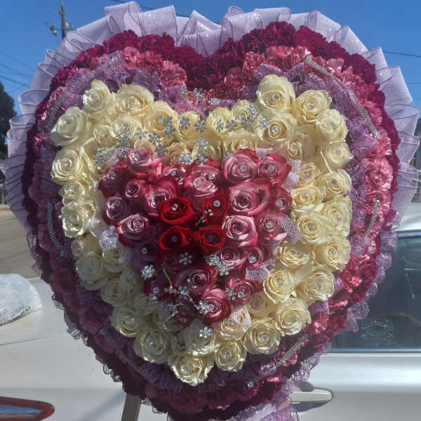 Heart-shaped rose arrangement in cream, pink, and red with purple ribbon trim
