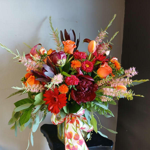 Mixed bouquet of orange, red, and pink flowers in a glass vase with a patterned ribbon