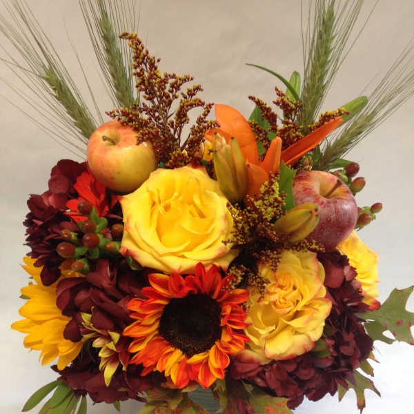 Mixed autumn bouquet with roses, sunflowers, apples, and lilies in a glass vase