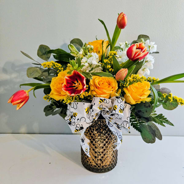 Yellow roses and tulips in a textured vase with a bee-pattern ribbon