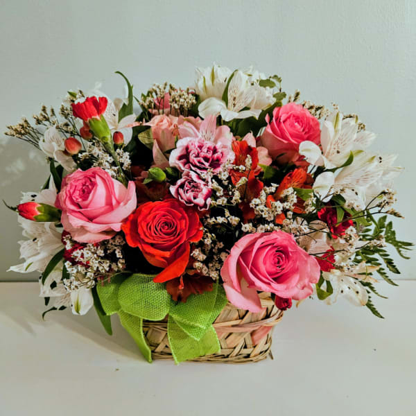 Mixed pink and red flower arrangement in a woven basket with a green ribbon