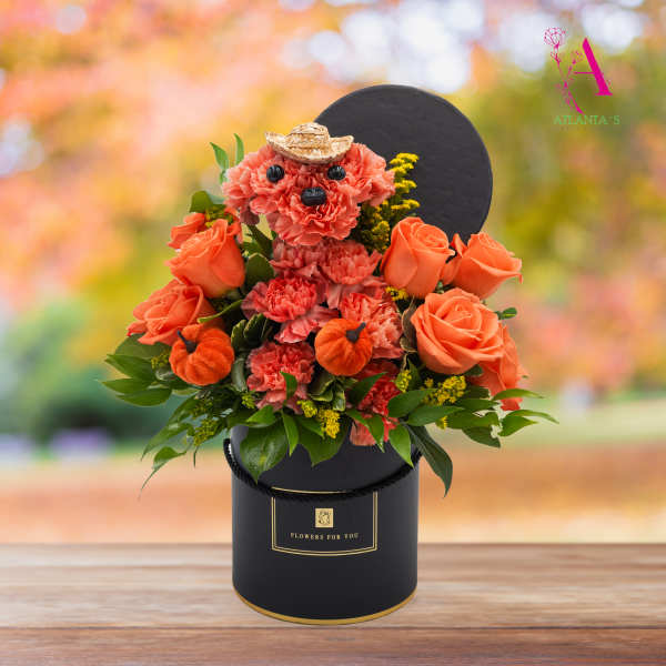 Orange rose and carnation arrangement in a black hatbox with a small straw hat accent