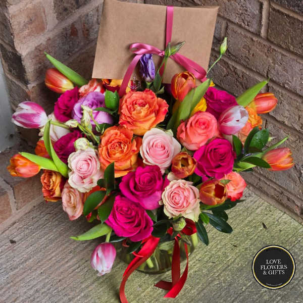 Bright bouquet of pink roses and orange tulips in a glass vase with a kraft card tied with pink ribbon.