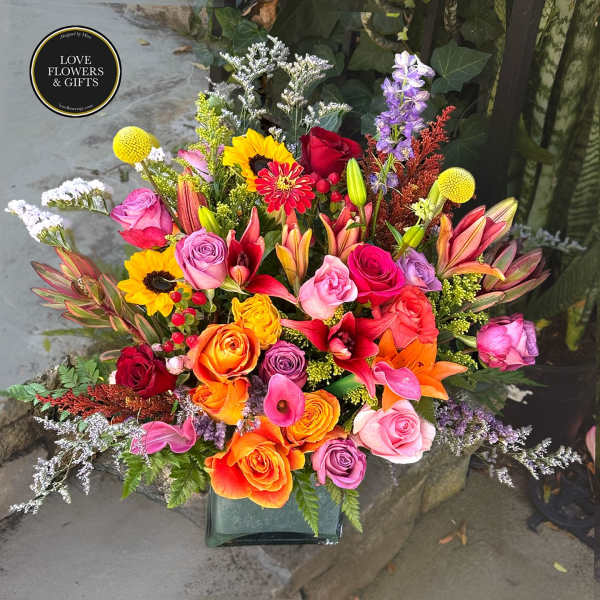 Vibrant mixed bouquet of roses, lilies, sunflowers, and other blooms in a square glass vase