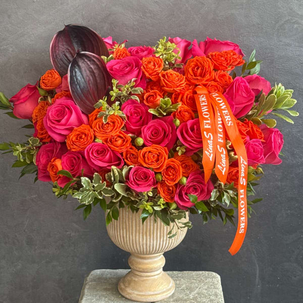 Pink and orange rose arrangement in a pedestal vase with dark decorative pods