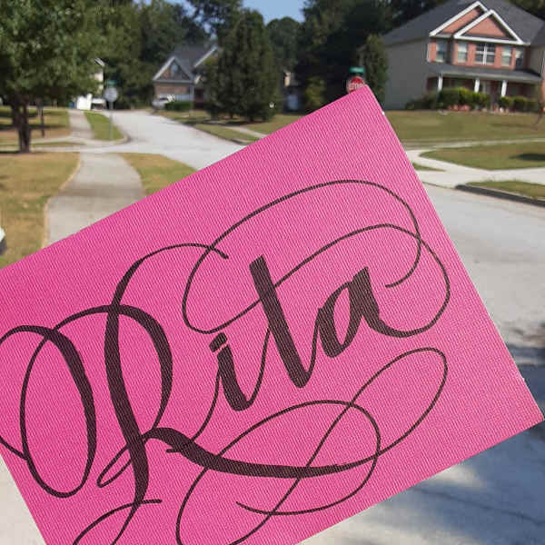 A pink sign with the name "Rita" held outdoors on a suburban street.