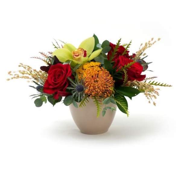 Low arrangement of red roses, green cymbidium orchid, and orange protea in a beige bowl vase