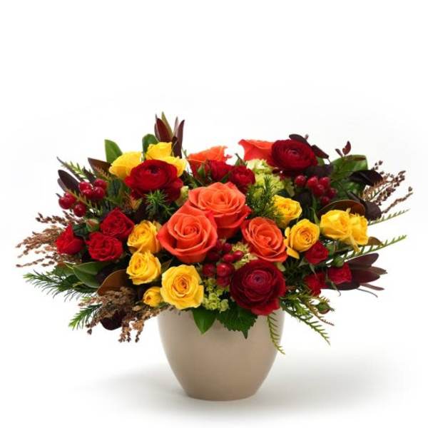Low arrangement of red, orange, and yellow roses with ranunculus in a beige ceramic vase.