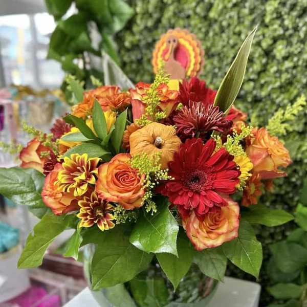 Fall arrangement of orange roses and red gerbera daisies in a round glass vase with a turkey pick