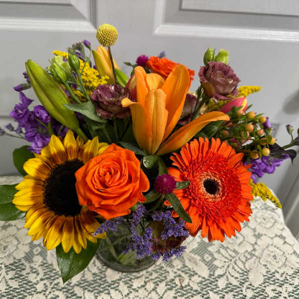 Mixed bouquet with sunflowers, orange roses, lilies, and gerbera daisies in a glass vase