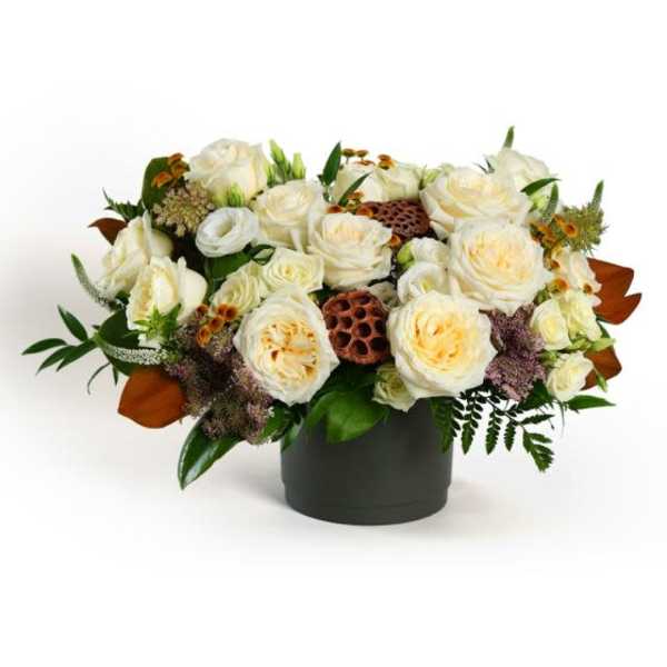 Low arrangement of cream roses with brown seed pods in a dark cylinder vase