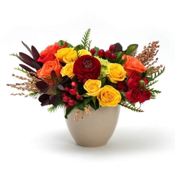 Low arrangement of yellow, orange, and red roses with berries in a beige vase