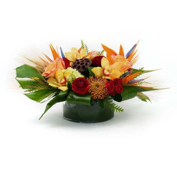 Low tropical arrangement of orange, yellow, and red flowers in a round glass vase