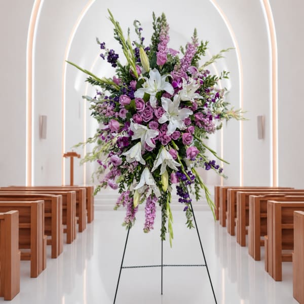 Tall floral spray with white lilies and purple blooms on a stand in a church aisle
