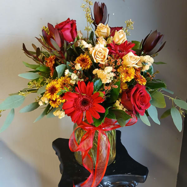 Mixed bouquet of red, yellow, and cream flowers in a glass vase with a red ribbon