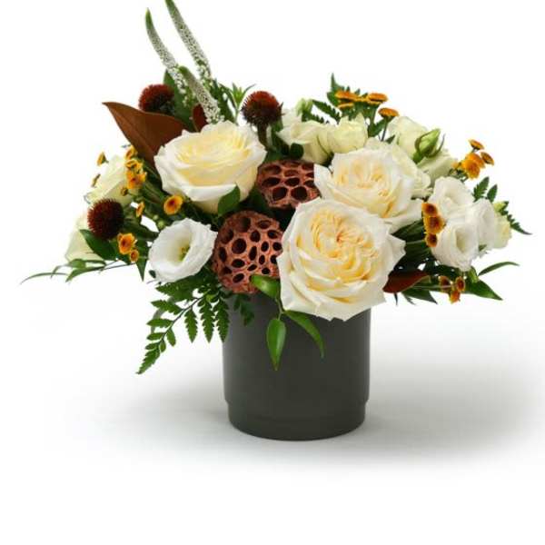Low arrangement of cream roses, white blooms, and yellow accents in a dark cylinder vase with lotus pods.