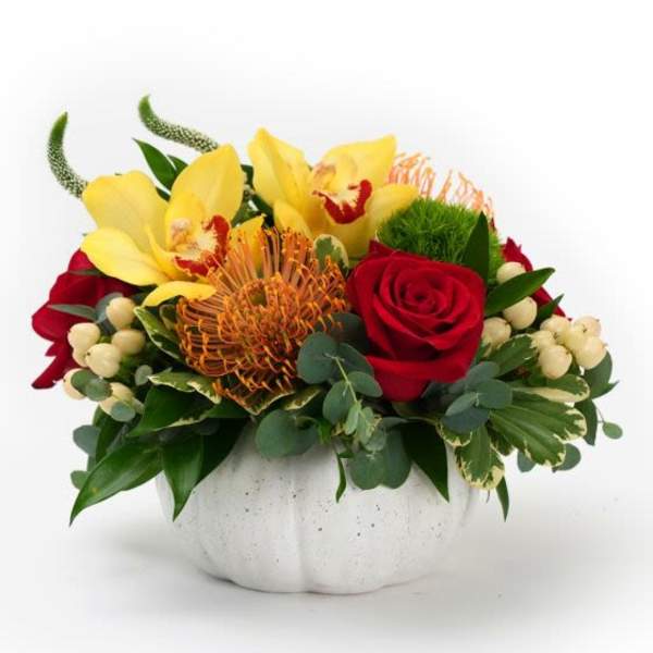 Low arrangement of yellow orchids, orange protea, and red roses in a white pumpkin-shaped bowl