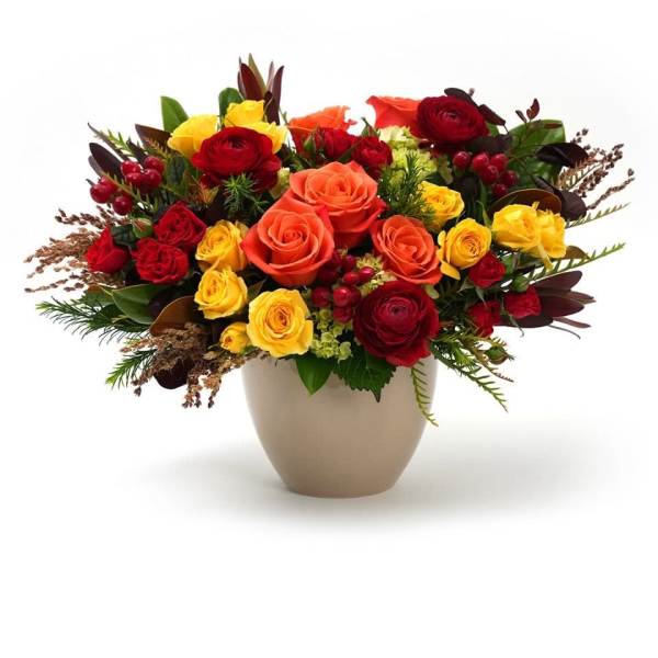 Low arrangement of orange, red, and yellow roses in a beige ceramic vase with red berries