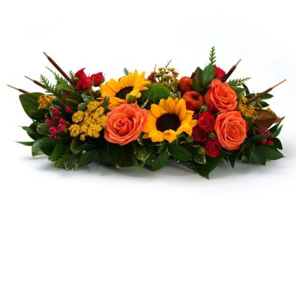 Low centerpiece of orange roses, yellow sunflowers, and red accent blooms in an elongated design