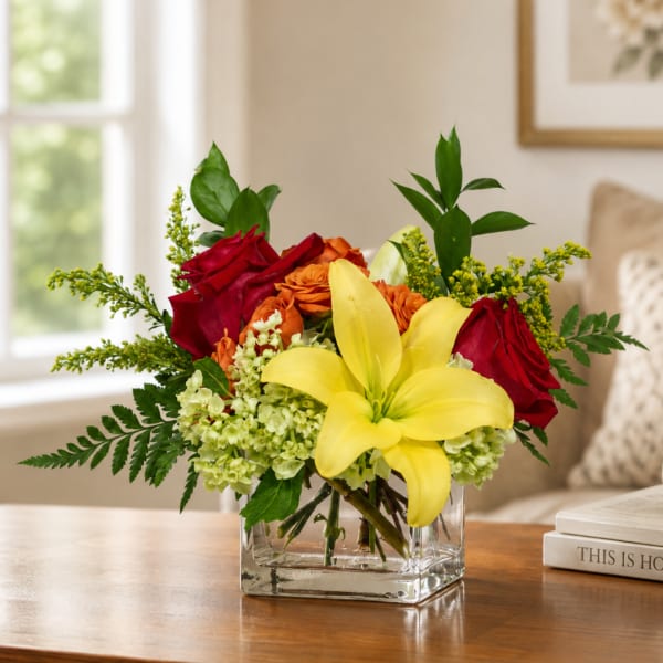 Mixed bouquet with yellow lilies, red roses, and orange flowers in a clear square vase