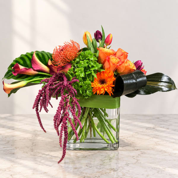 Colorful bouquet in a clear square vase with roses, calla lilies, and orange blooms