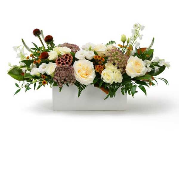 Low arrangement of cream roses with orange accents and seed pods in a white rectangular container
