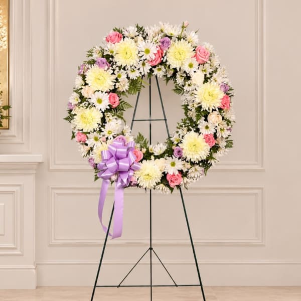 Round floral wreath of white, pink, and lavender flowers on a stand with a purple bow
