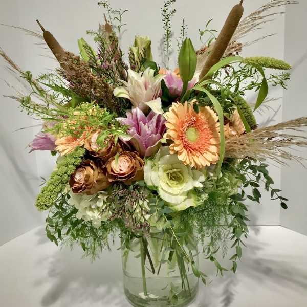 Tall rustic bouquet of peach gerbera, roses, lilies and white hydrangea in a glass vase with cattails and grasses.