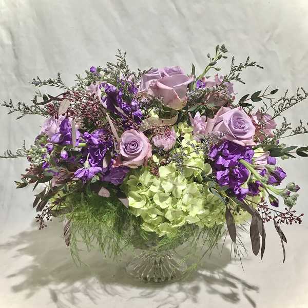 Low pedestal arrangement of lavender roses, purple blooms, and green hydrangeas in a glass compote vase