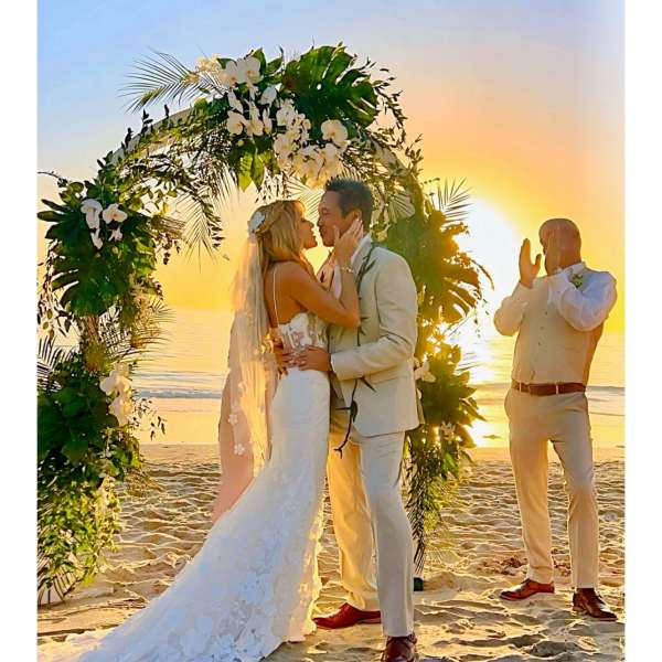 Bride and groom kissing under a floral arch on a beach at sunset