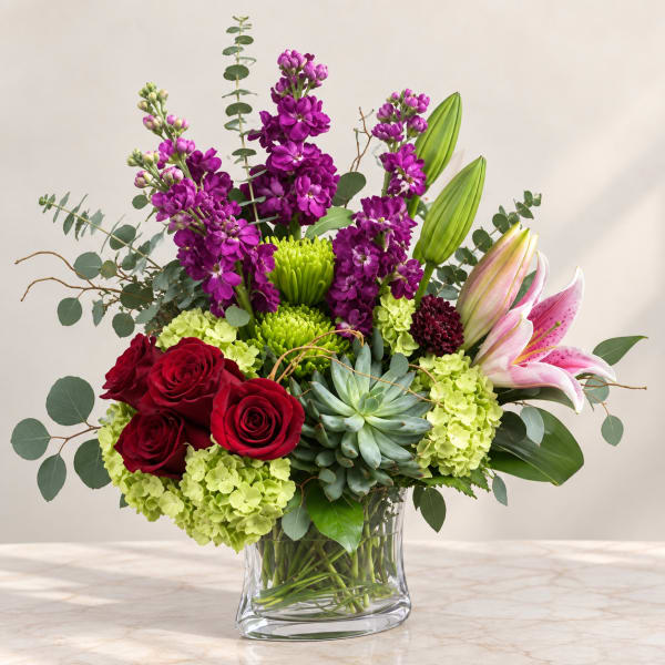 Mixed bouquet of red roses, purple blooms, lilies, and green hydrangeas in a glass vase