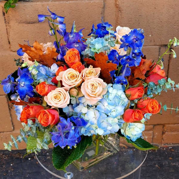 Arrangement of blue hydrangeas, blue delphinium, and orange and peach roses in a clear glass vase