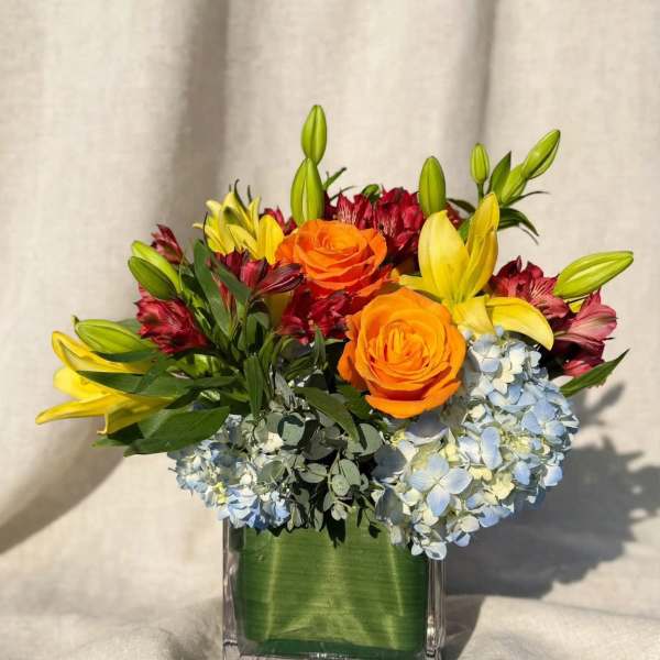 Orange roses and yellow lilies in a square glass vase with blue hydrangeas