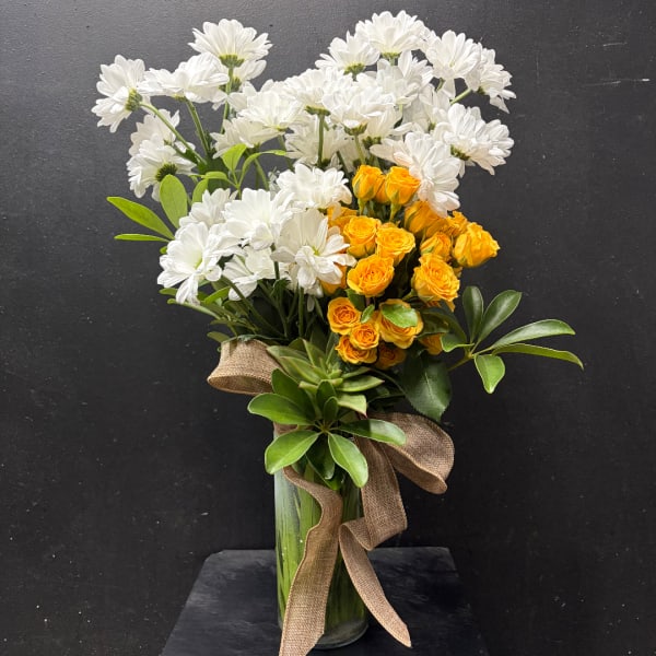 White daisies and yellow spray roses in a glass vase with a burlap ribbon