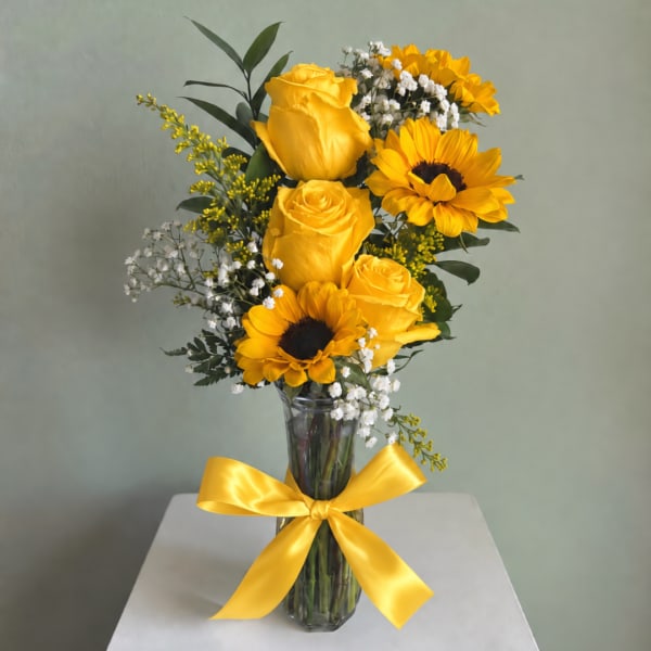 Yellow roses and sunflowers in a clear glass vase with a yellow ribbon