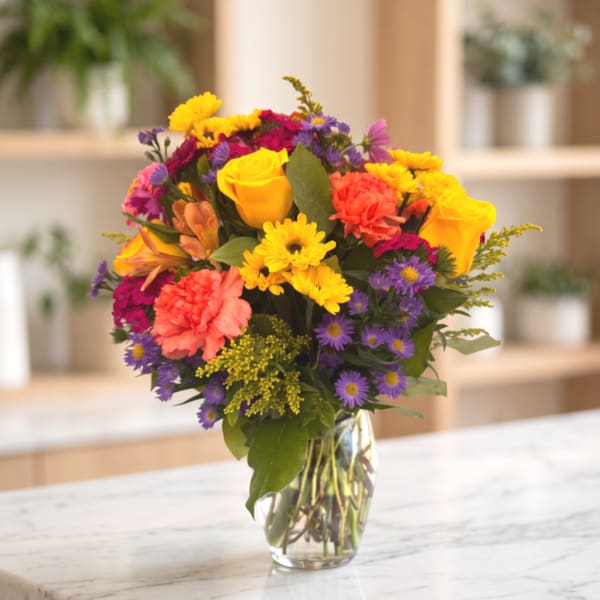Mixed bouquet of roses, carnations, and purple daisies in a clear glass vase
