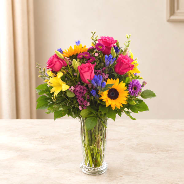Mixed bouquet of roses, sunflowers, lilies, and purple daisies in a clear glass vase