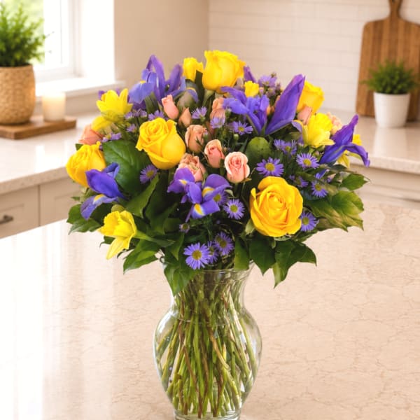 Bouquet of yellow roses, purple irises, and small pink flowers in a clear glass vase