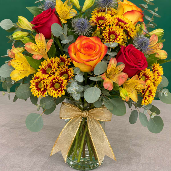 Mixed bouquet of roses, lilies, and chrysanthemums in a glass vase with a gold ribbon