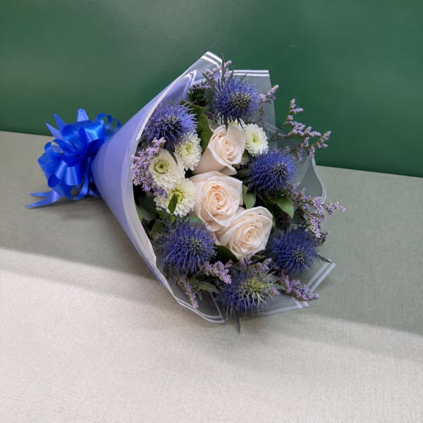 Bouquet of pale roses, white blooms, and blue thistle wrapped in lavender paper