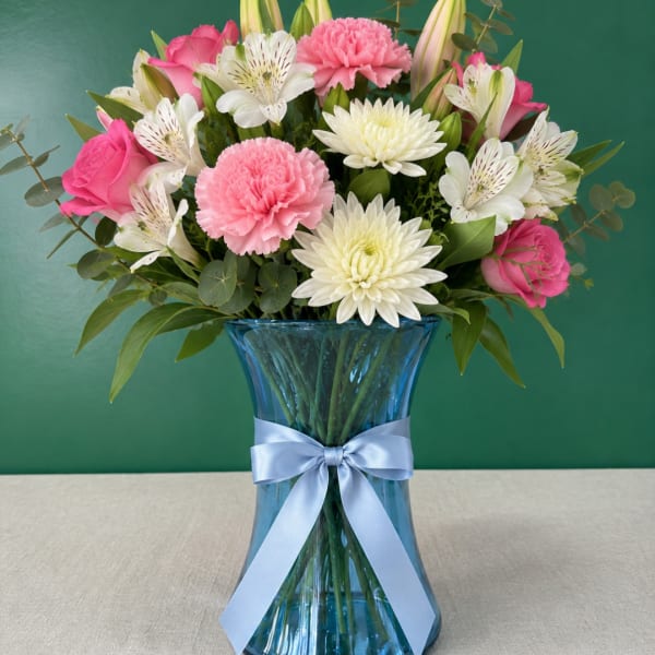 Pink roses, carnations, and white daisies in a blue glass vase with a ribbon